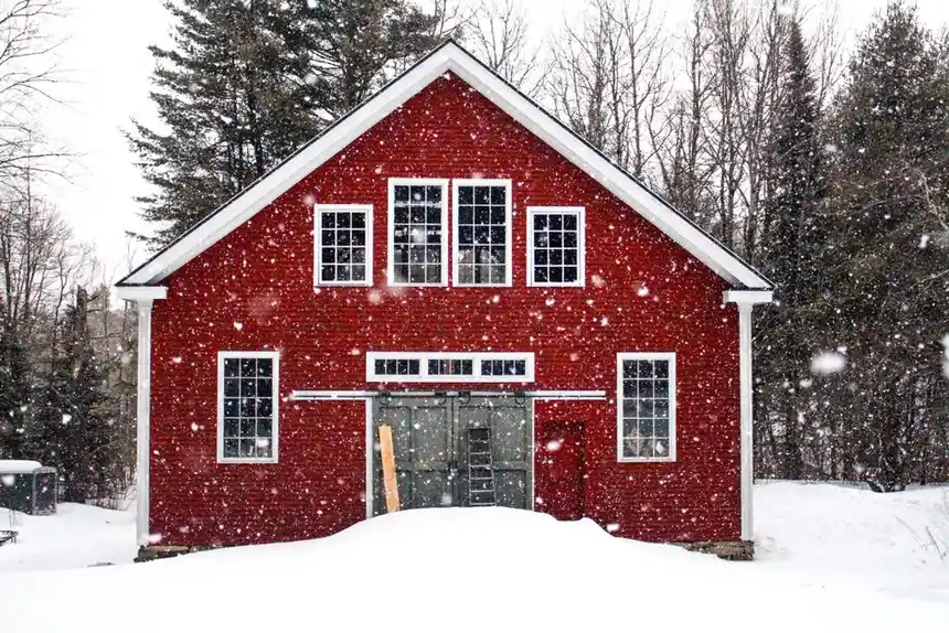 Charming red barn surrounded by falling snowflakes and leafless trees during a serene winter day.
